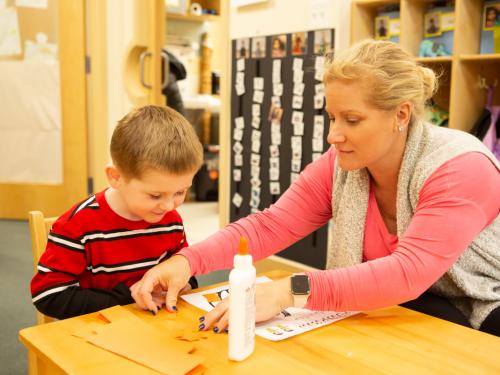 Female teacher works 1-on-1 with a young boy on an art project in a classroom.
