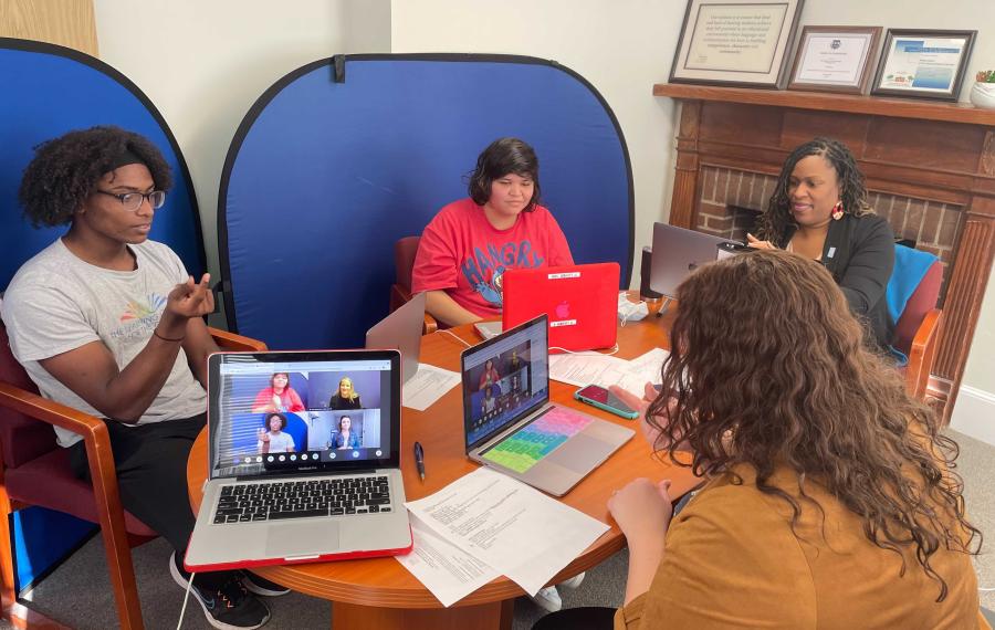 Two students facing the camera are sitting in front of laptops with blue screens in the background. 