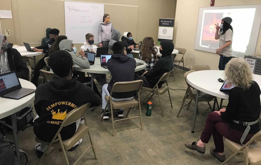 Students sit at round tables while a presenter stands in the front of the room.