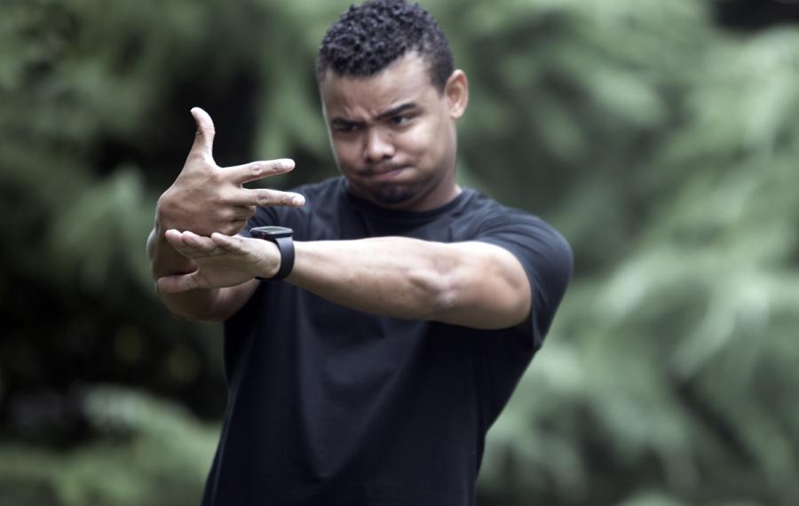 A man of color pictured outside wearing a black short sleeved shirt. Both hands are outstretched in front of him.