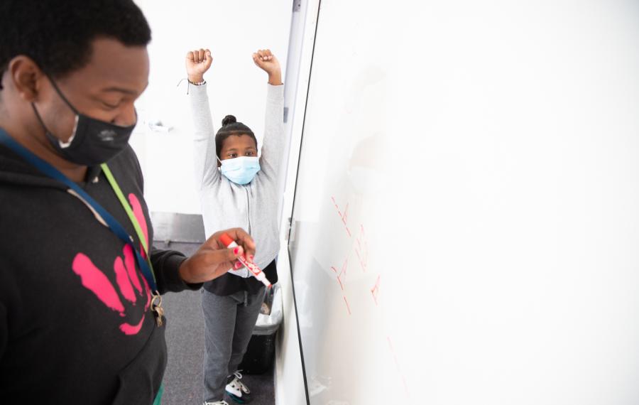 A teacher wearing a face mask is writing on a white board. In the background, a student raises his hands over his head in celebration.