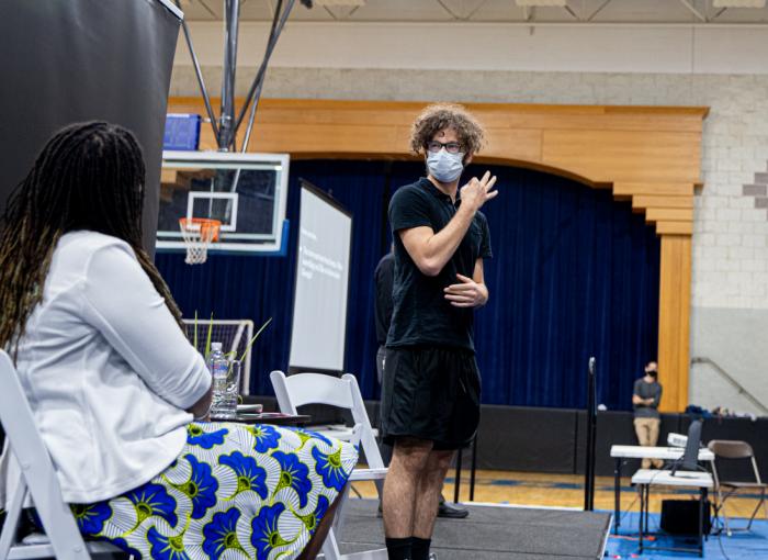 Left: Glennis Matthews sits on a chair. Right, a male student wearing a face covering faces her.