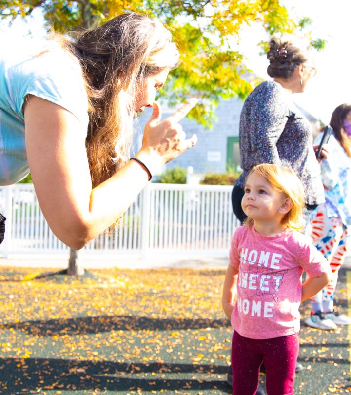A woman standing on the left with long blonde hair is signing to a young girl with dark hair wearing a pink shirt.