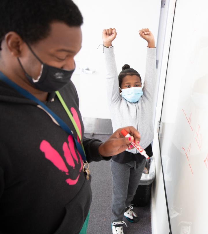 A teacher wearing a face mask is writing on a white board. In the background, a student raises his hands over his head in celebration.