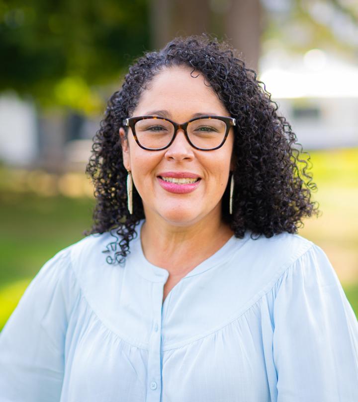 A Latinx woman standing outside. Curly black hair and glasses and a light blue shirt.