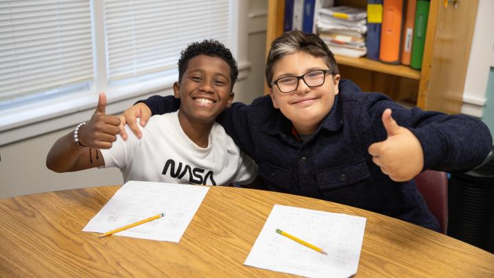 Two young bpys are sitting side by side at a desk. They are both smiling at the camera, Each has a thumbs up sign.