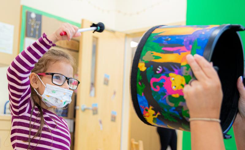 A young student with glasses and a face mask is about to bang on a drum being held by their teacher.