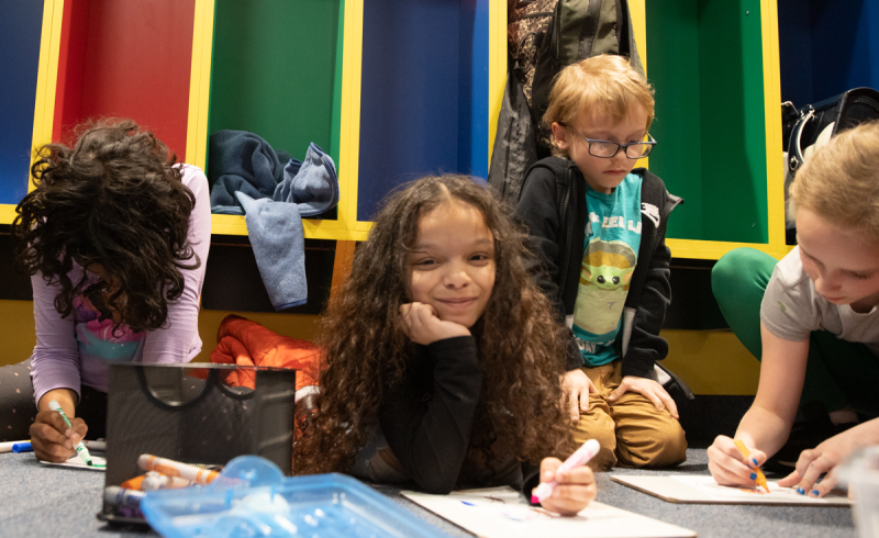Four students sitting on the floor doing an art project. The second girl from left is looking up and smiling.