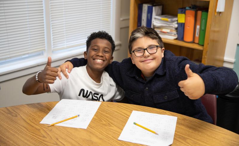 Two young bpys are sitting side by side at a desk. They are both smiling at the camera, Each has a thumbs up sign.
