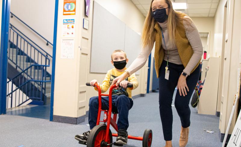 A female wearing a face mask in a school hallway. To the left, a young student riding a tricycle.
