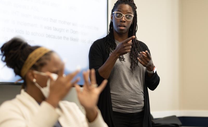 A female Black Deaf Interpreter is focused in the background. 