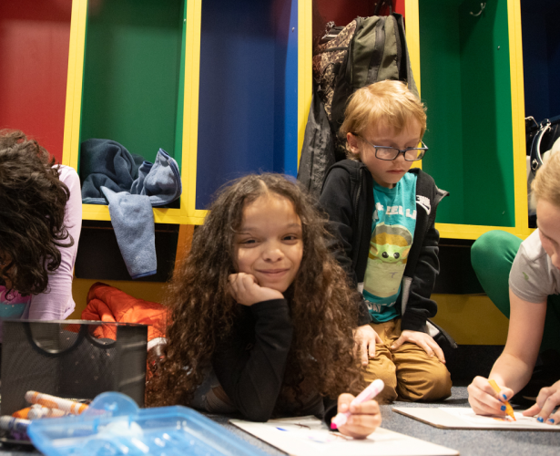 Four students sitting on the floor doing an art project. The second girl from left is looking up and smiling.