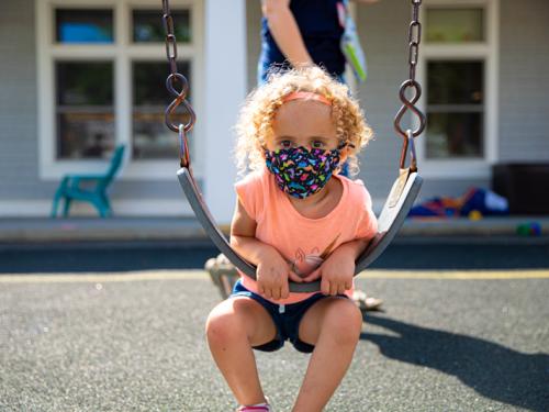 A young student with blonde curly hair wearing a pink shirt sitting on a swing.