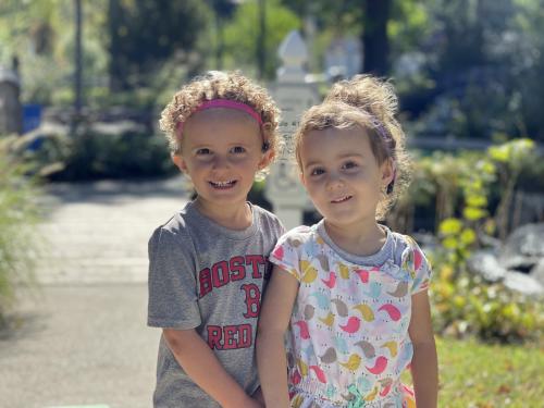 Two young students are smiling side by side standing outside.