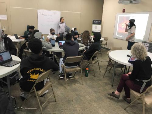 Students sit at round tables while a presenter stands in the front of the room.