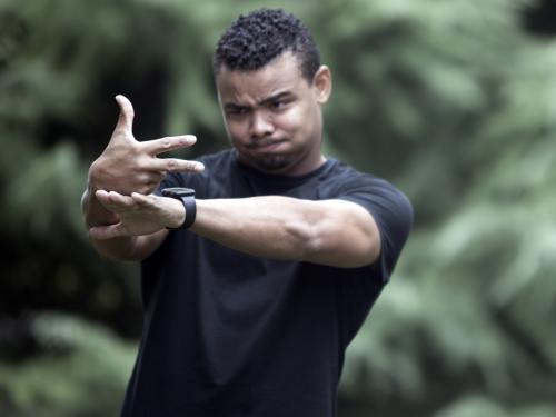 A man of color pictured outside wearing a black short sleeved shirt. Both hands are outstretched in front of him.