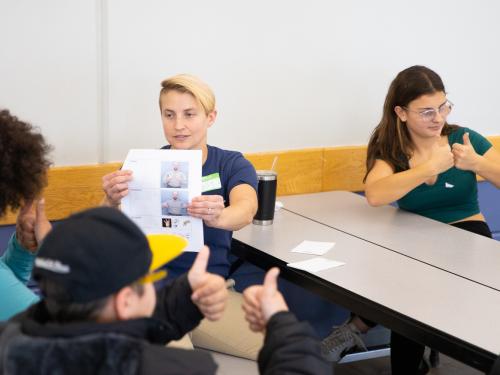 A group of students sitting at a long table. In focus, a young female offering a sign in ASL. Another individual is holding up a paper with an ASL sign on it. Blurred in the foreground are two students not facing the camera holding two thumbs up signs.