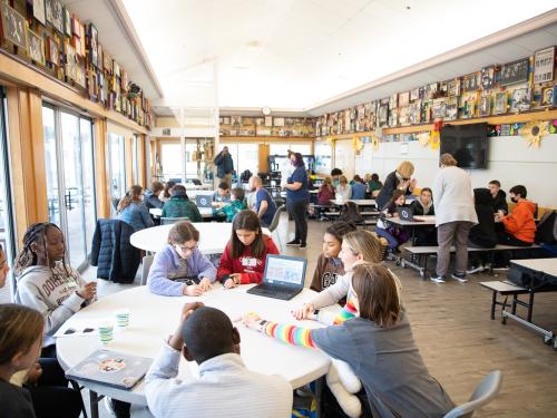 A large group of students seen sitting at various tables in a cafeteria.