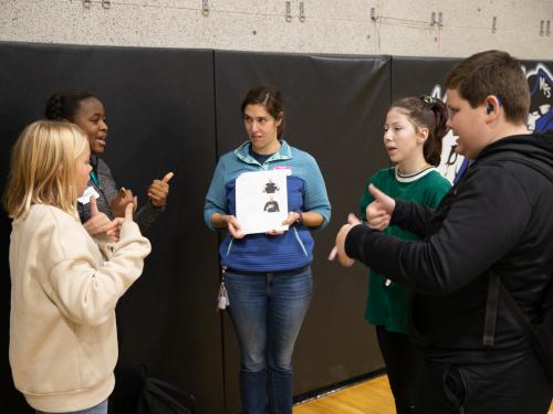 A group of students standing in a circle. In the middle, a woman is holding a piece of paper with an ASL sign printed. Two groups of students are showing that sign to each other.