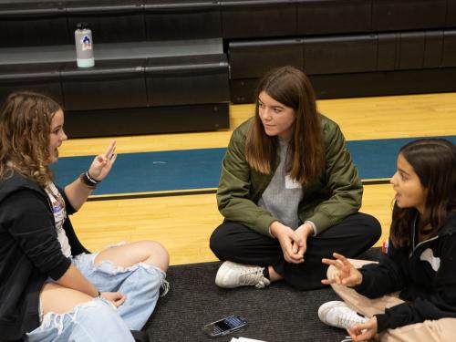 Three female students sitting in a circle on the gym floor. They are teaching each other ASL signs.