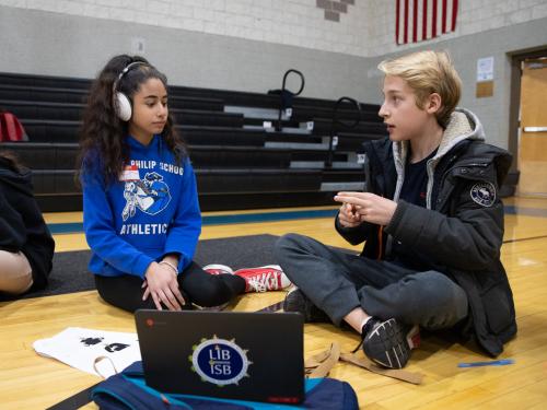 Two students sitting on the gym floor. On the left is a female student wearing a blue MPS ghosts sweatshirt, and is wearing earphones. On the right, a young student in a black jacket is communicating in ASL.