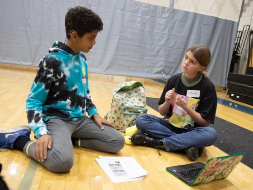 Two students sitting on the gym floor looking at each other and communicating. On the left, a young male student in a tye-dye sweatshirt. On the right, a young female student.