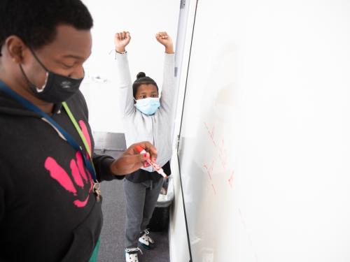 A teacher wearing a face mask is writing on a white board. In the background, a student raises his hands over his head in celebration.