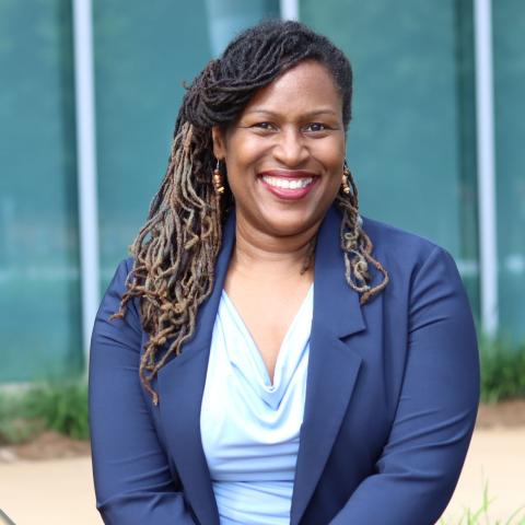 A black woman with long dark hair sitting outside. She is wearing a dark blue blazer over a light blue shirt and smiling.