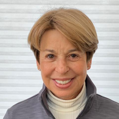 A white woman with short blonde hair and gray jacket. White wall in background.