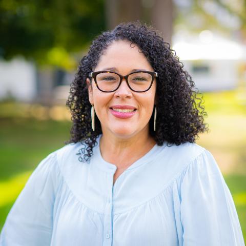 A Latinx woman standing outside. Curly black hair and glasses and a light blue shirt.