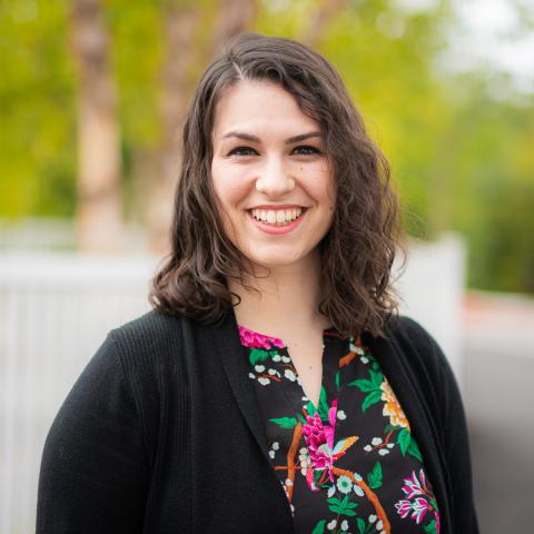 A white woman standing outside with brown hair. She is wearing a floral shirt under a black cardigan.