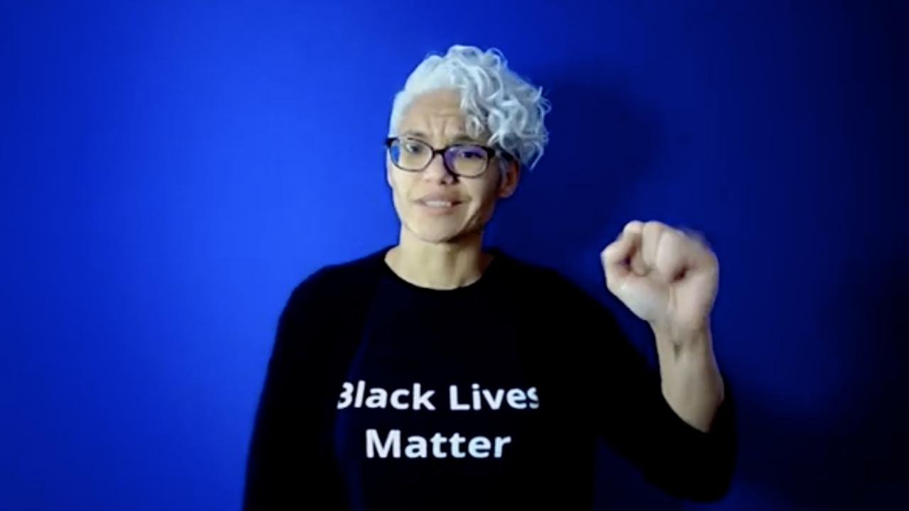 A woman with short white curly hair and glasses stands in front of a blue background. She is wearing a black shirt with white letters BLACK LIVES MATTER